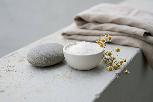 Smooth river stone, bath salt bowl, and dried chamomile on grey concrete in calm diffused daylight.