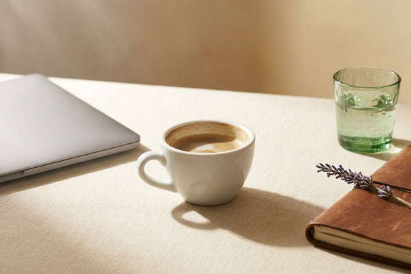 Closed laptop, half-empty espresso, water glass, and lavender on a cream linen desk in soft midday light, evoking the post-lunch afternoon dip.