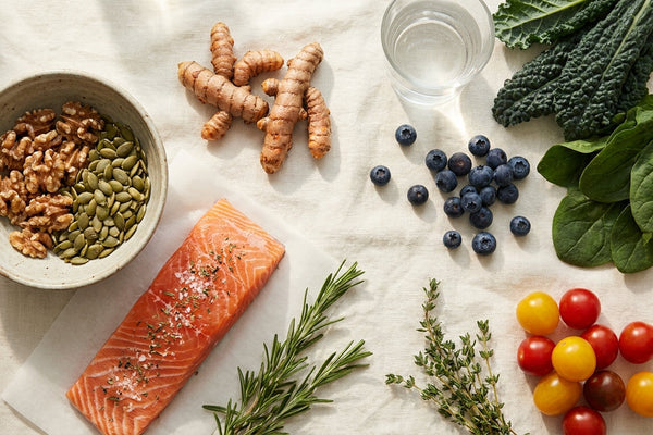 Editorial flatlay of anti-inflammatory whole foods on cream linen: salmon, turmeric, walnuts, pumpkin seeds, leafy greens, blueberries