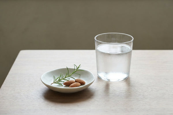 Minimalist still life on walnut wood: a glass of water, a small ceramic dish, and a leaf sprig in soft daylight