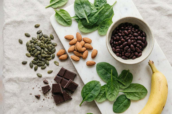 Bright overhead flatlay of magnesium-rich foods on cream linen and white marble: pumpkin seeds, almonds, dark chocolate, spinach, black beans, and a banana