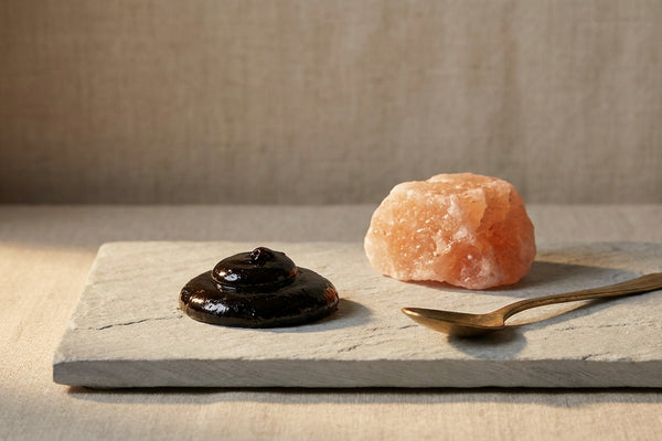 Dark shilajit resin on light grey slate beside a piece of himalayan rock and a small brass spoon in warm side light